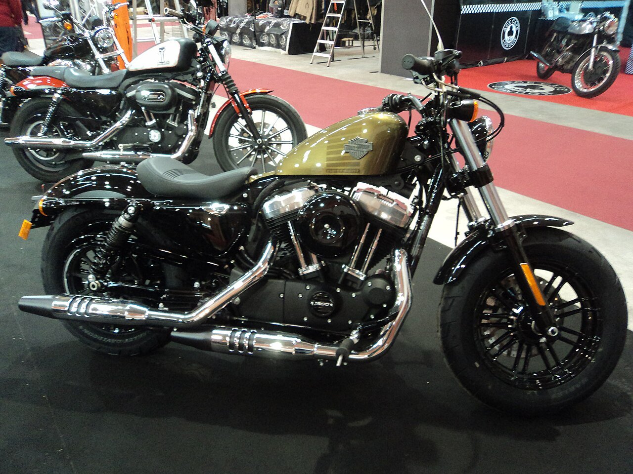 אופנוע Harley-Davidson בצבע חום-שחור עם מנוע 1200, צילום צד בקרבת אופנועים נוספים על שטיח אדום בפנימיות תערוכה.