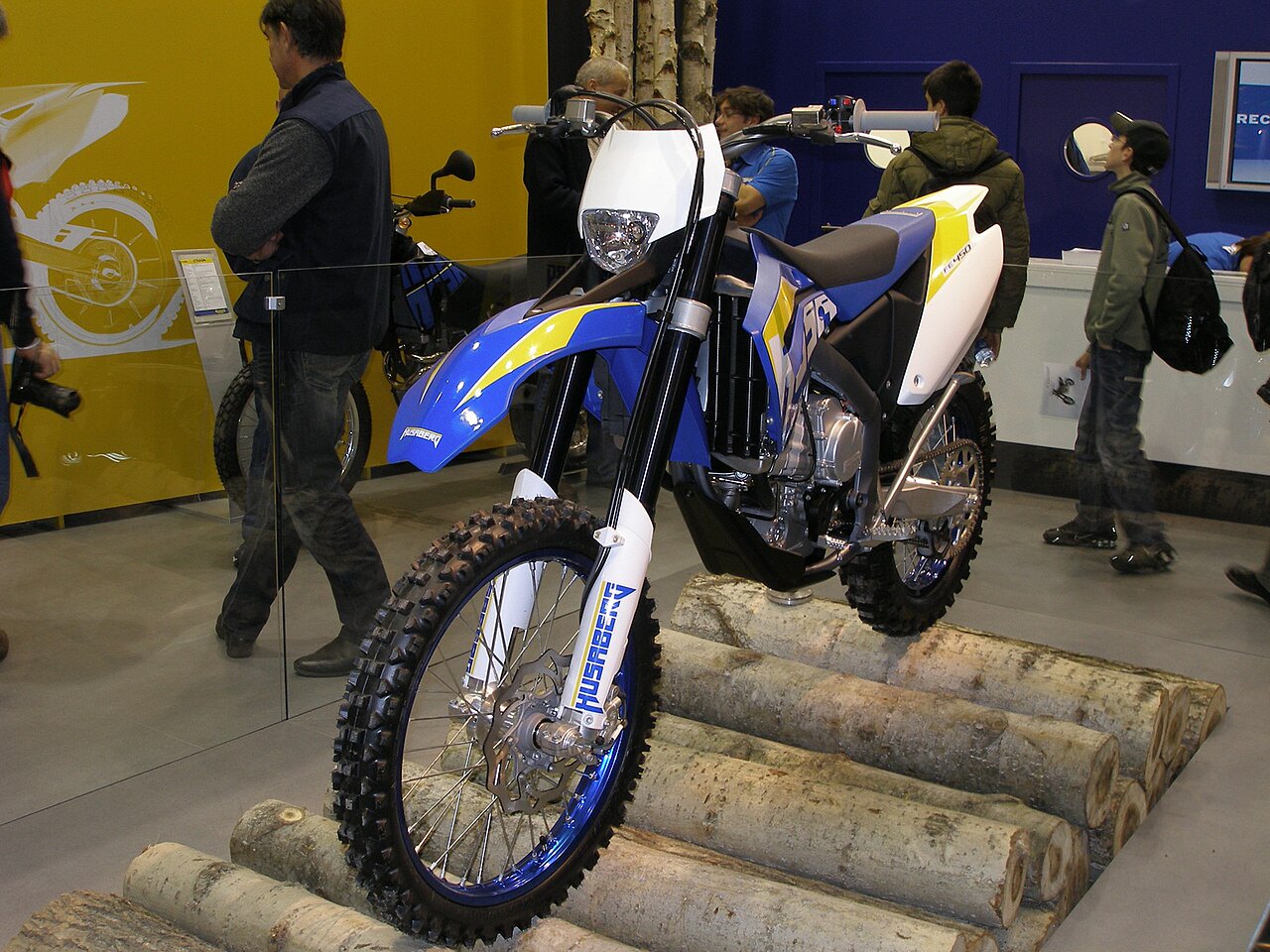 אופנוע Husqvarna FC 450 בצבעי כחול, צהוב ולבן, מוצג על עצי בורש בזווית קדמית, עם מבקרים ברקע בפנימיות תערוכה.