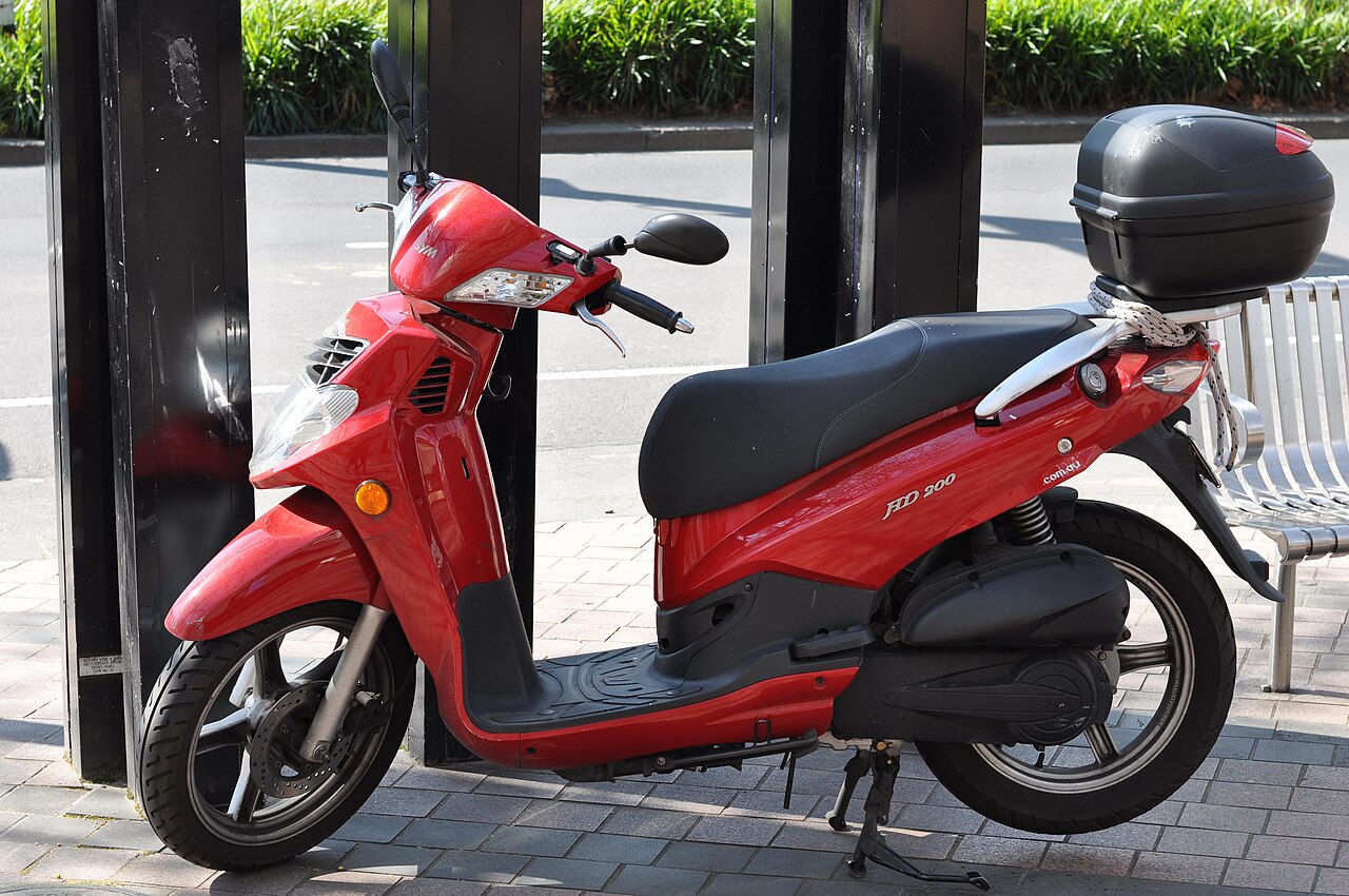 אופנוע חשמלי אדום מדגם HD 200 של חברת contact, מונח על מדרכה בזווית צדית, עם מזוודה שחורה אחורי, בסביבה עירונית עם שיחים