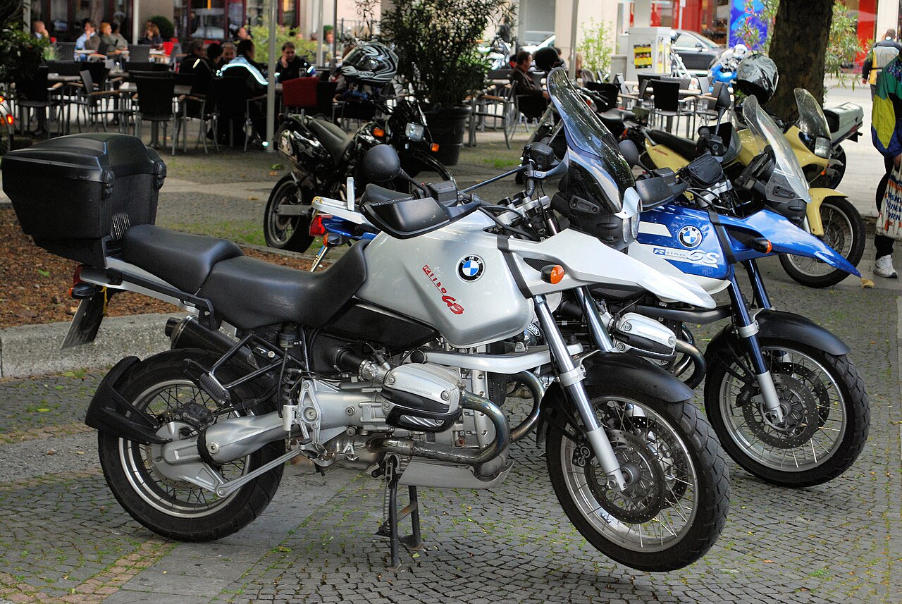 אופנוע BMW R1150GS כסף עם מזוודה אחורייה, מונח על רצפת אבן מול מסעדה חיצונית עם אנשים יושבים בשולחנות
