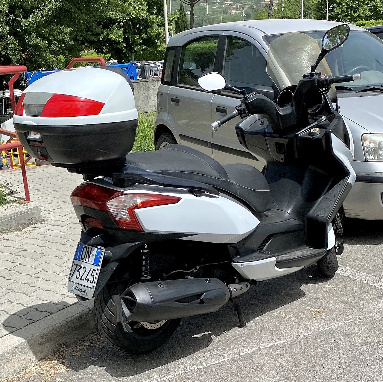 אופנוע סקוטר לבן-שחור מותג Yamaha, זווית אחורי-צד, עם מזוודה על המושב, חנייה בחצר עם מכונית אופל סילבר ברקע.