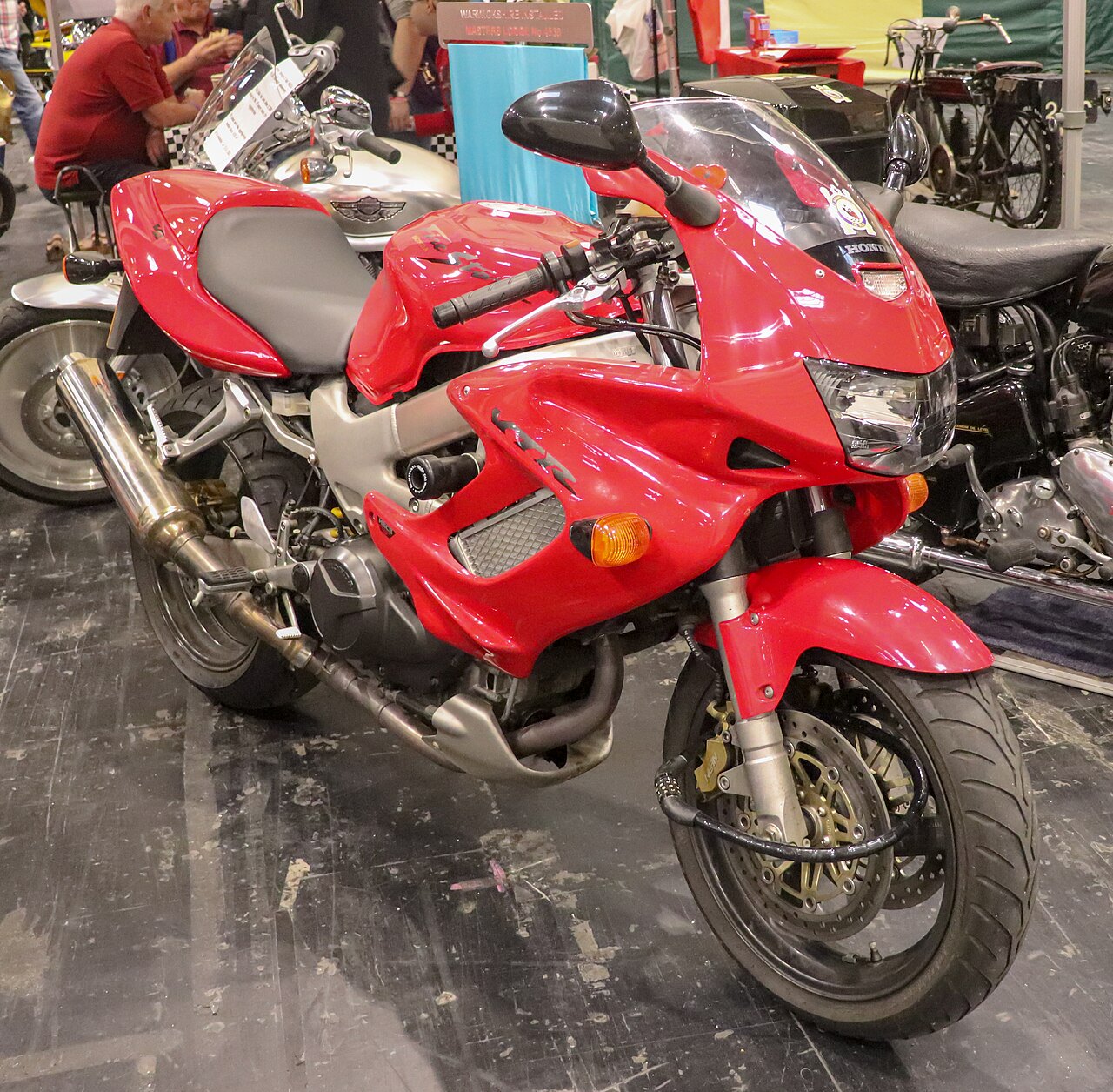 אופנוע הונדה VFR אדום בהיר, צילום קרוב מזווית צד-קדימה, מוצג בתערוכה עם אופנועים נוספים ברקע.
