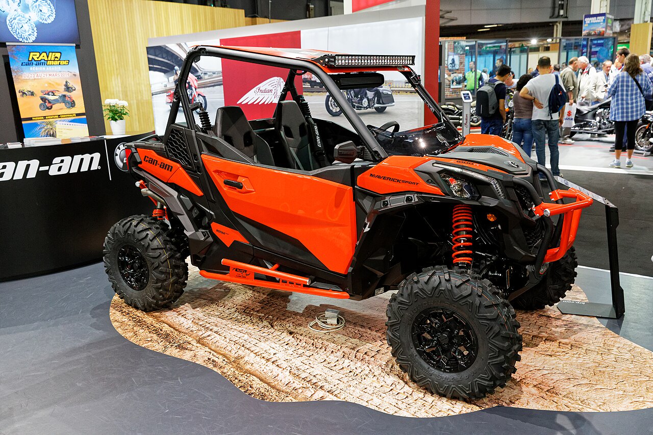רוכב אופנועי שטח Can-Am Maverick X3 צבע כתום-שחור, מוצג בתערוכה עם גלגלים גדולים ומערכת תאורה עליונה, ברקע תצוגות ומבקרים.