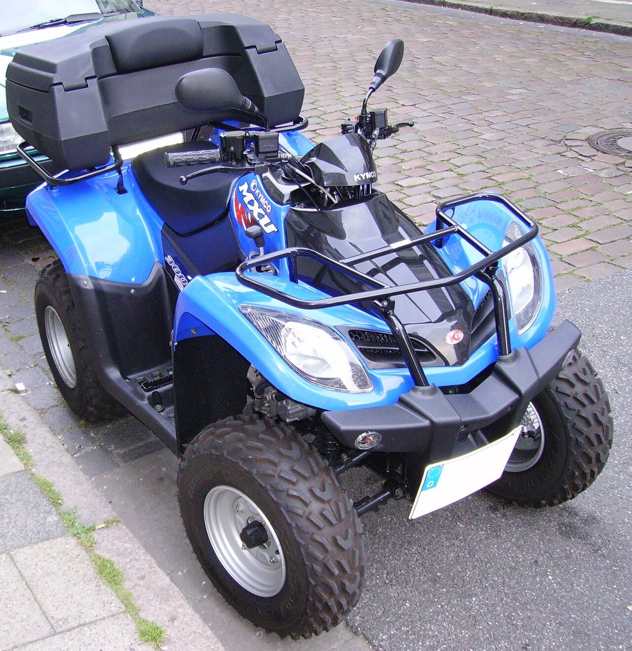 אופנוע ארבעה גלגלים KYMCO MXU 500 בצבע כחול בהיר, צילום מזווית עליונה, עמיד על רחוב אספלט עם פלטפורמות אבן, עם מארז אחורי