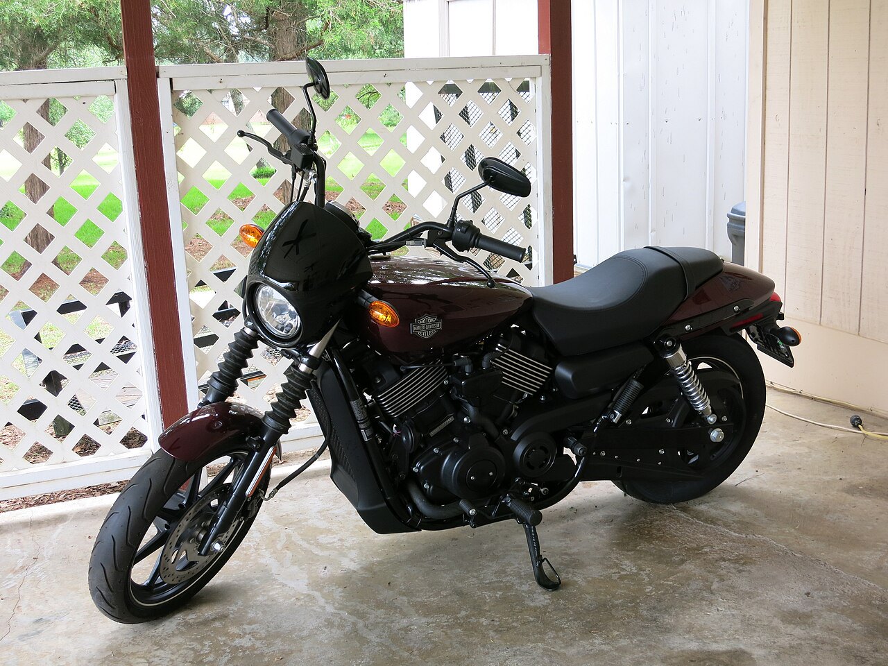 אופנוע Harley-Davidson בצבע חום-שחור, צילום צד, על סלון חיצוני עם גדר לבנה וירוק של גן ברקע
