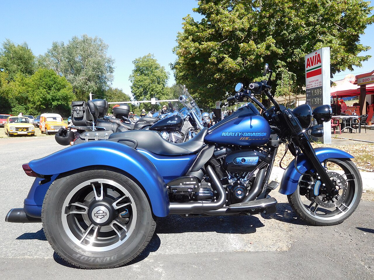 אופנוע תלת-אופן Harley-Davidson 114 כחול בהיר, צילום צד, חנייה חיצונית עם עצי רקע ותחנת דלק AVIA