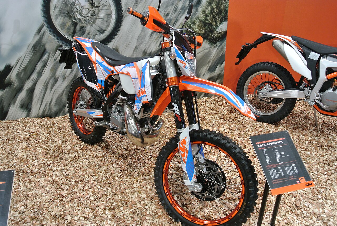 אופנוע KTM 200 EXC & POWERPARTS בצבעי כתום, לבן וסגול, מוצב על שכבת עלים יבשים, עם שלט תכונות ורקע של אופנוע בפעולה.