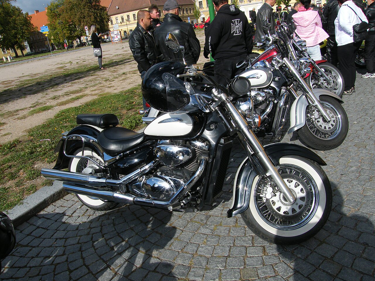 אופנוע Harley-Davidson קלאסי בצבע שחור-לבן, צילום מזווית צד קרוב על רצפת אבן, עם קבוצת אנשים ואופנועים נוספים ברקע.
