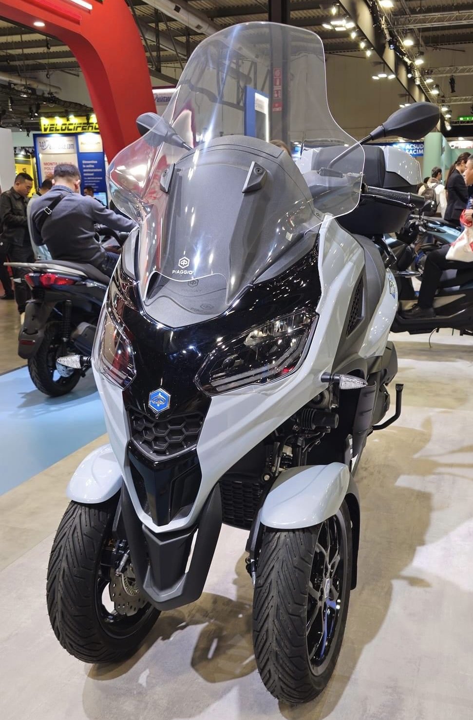 אופנוע תלת-גלגלי Piaggio MP3 לבן-שחור בתצוגה, זווית קדמית עם מראה ומסך עליון, במعرض רכבים עם מבקרים ברקע