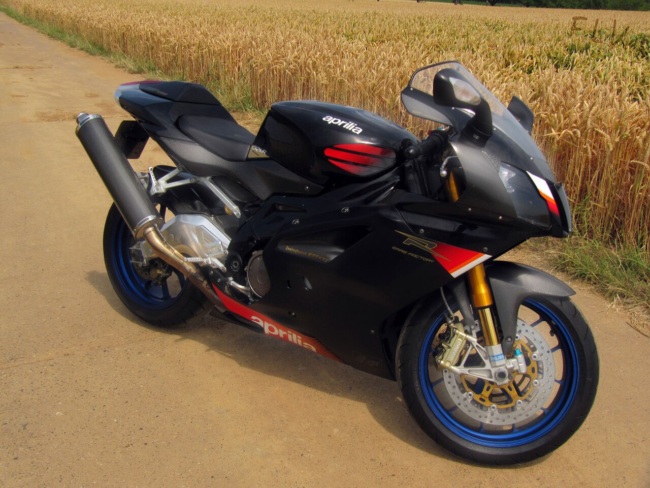 אופנוע Aprilia RSV4 שחור עם פסים אדומים, צילום צד מימין על דרך עפר מול שדה חיטה, גלגלים כחולים ומערכת תיבת גלגלים זהובה