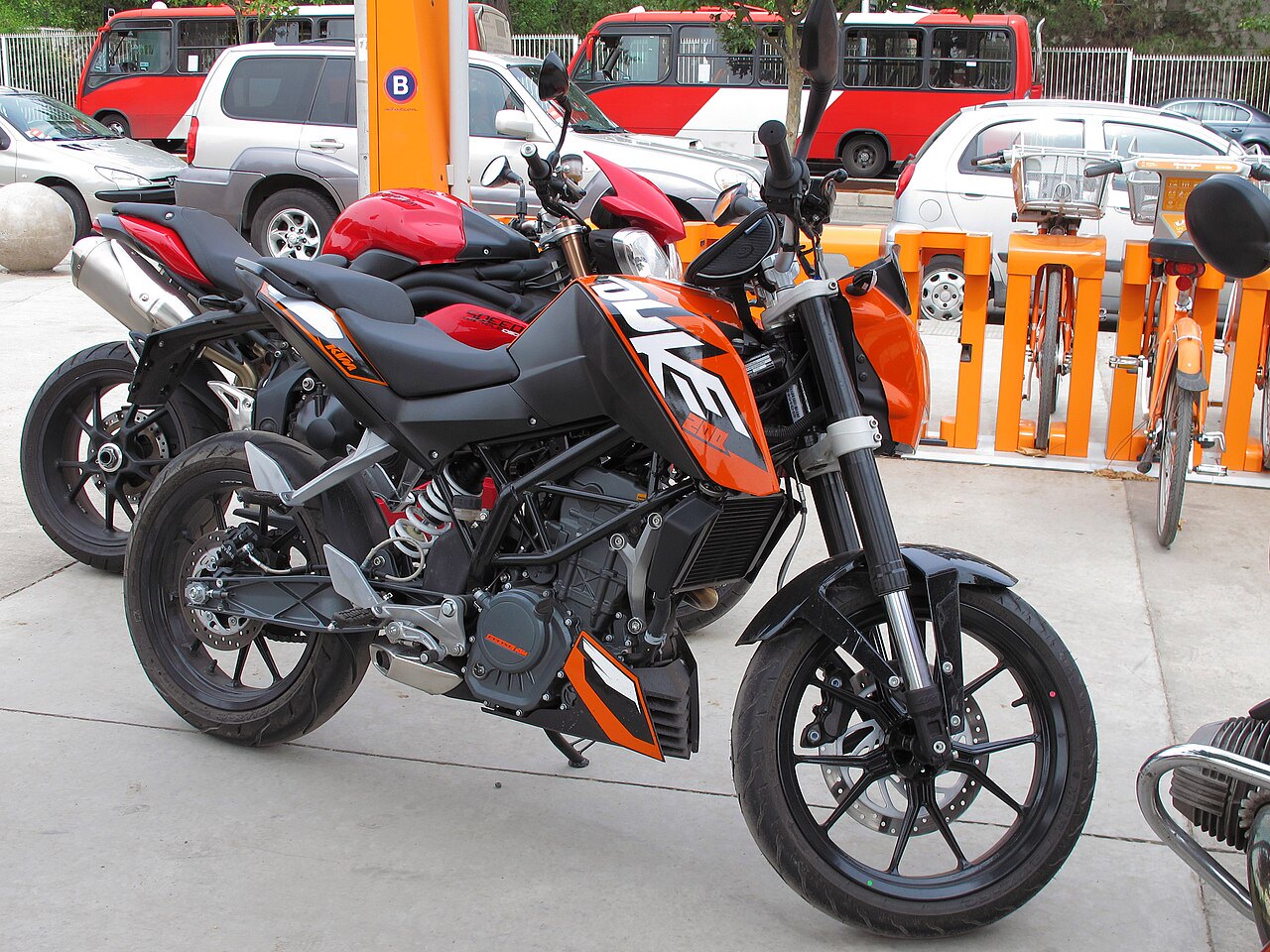אופנוע KTM Duke 390 בצבע כתום-שחור, צילום קרוב מזווית צד, עמיד על המדרכה עם אופנוע אדום ומכוניות ברקע