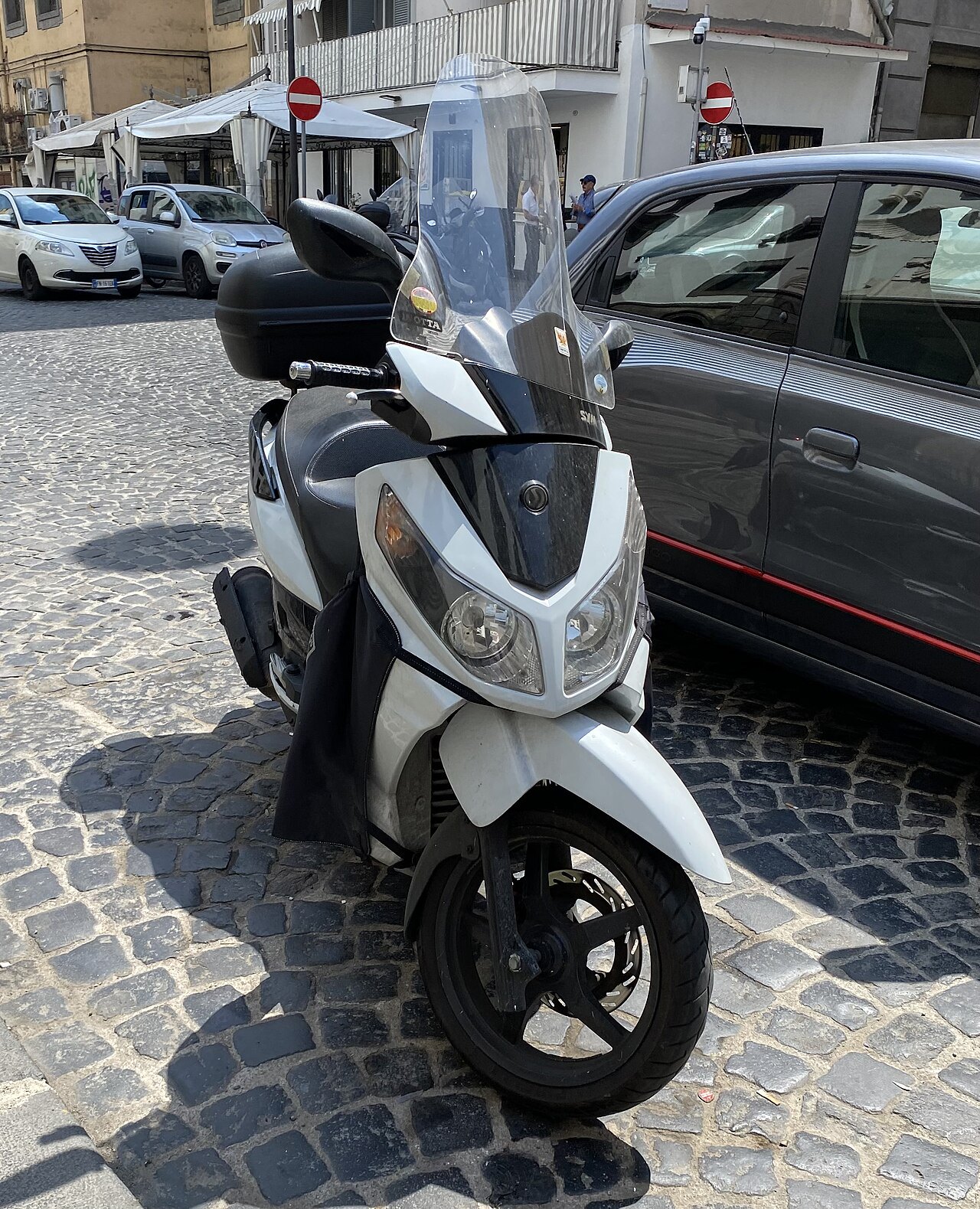 אופנוע סקוטר לבן-שחור של סוזוקי, מותקן עם מוגן זכוכית וקופסה אחורייה, עמד על רחוב אבן עם מכוניות בסביבה