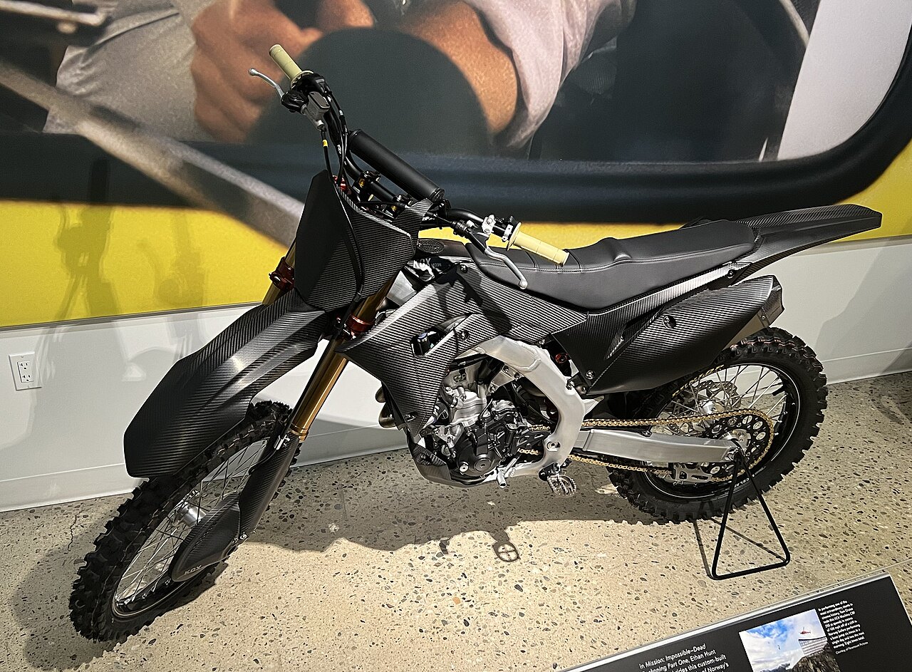 אופנוע מוטו-קרוס מותג KTM בצבע שחור עם פלסטיק פחמן, צולם בזווית צדית על רקע קיר צהוב עם ציור של רוכב, בתצוגה פנימית.