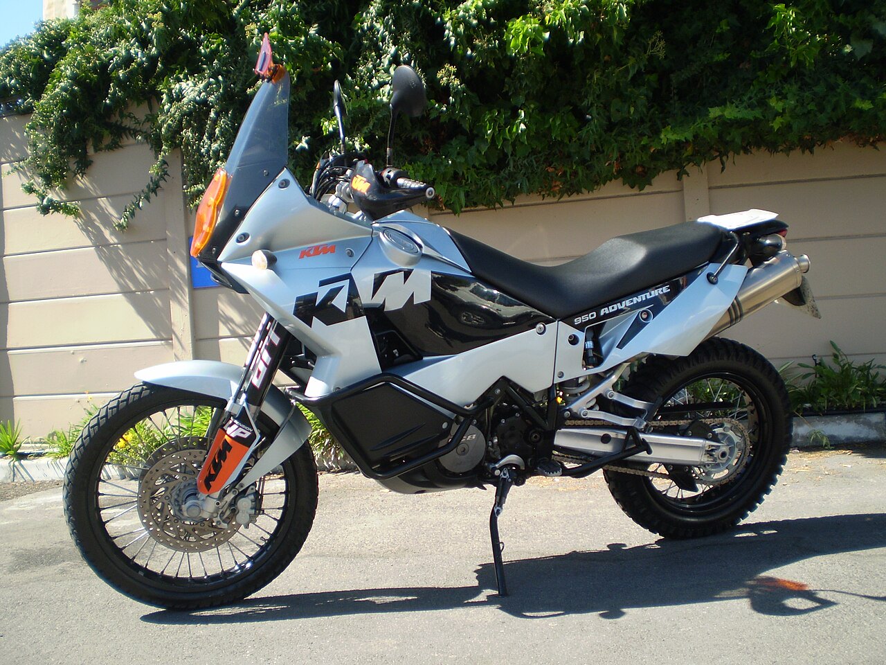 אופנוע KTM 950 Adventure לבן-שחור, צילום צד, על אספלט מול חומה עם צמחייה ירוקה