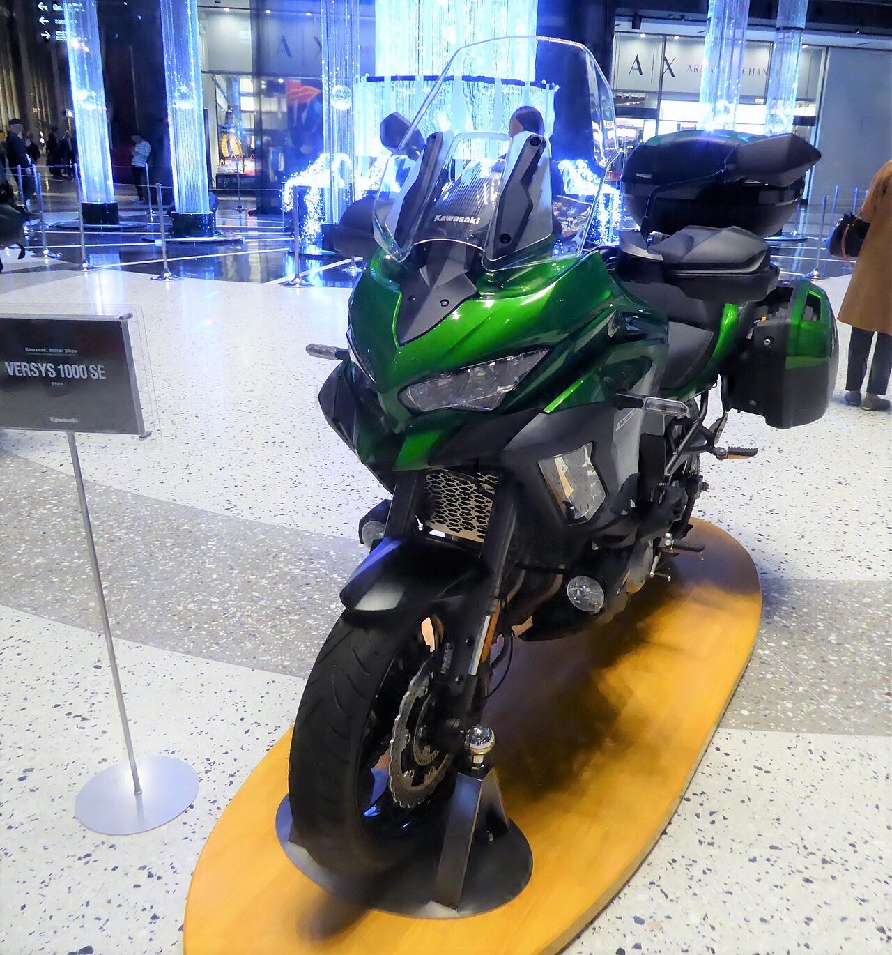 אופנוע Kawasaki VERSYS 1000 SE בצבע ירוק בהיר, צולם מזווית קדמית-צדית על פלטפורמה עץ בפנימיות מרכז קניות מואר, עם שלט תיאור