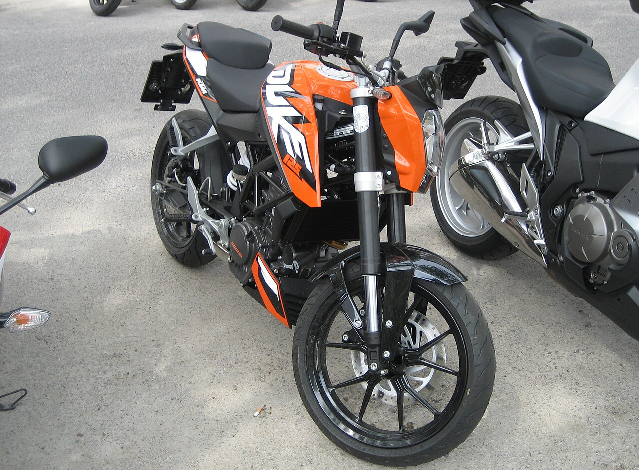אופנוע KTM Duke 390 בצבע כתום-שחור, צילום קרוב מזווית עליונה, עמיד על אספלט, לצד אופנועים אחרים
