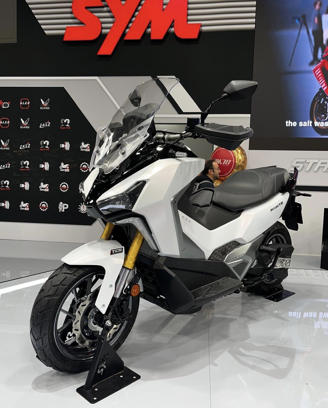 אופנוע SYM M12T6 לבן-שחור בתצוגה, זווית צד, עם מראה עליון וסיבוב קדמי, ברקע לוגו SYM וסמלים של טכנולוגיות.