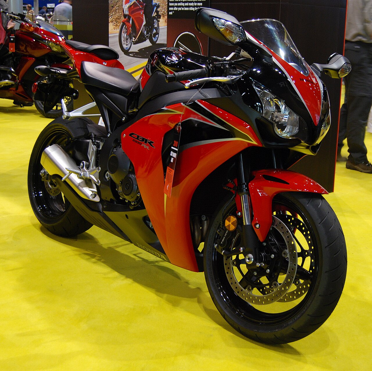 אופנוע הונדה CBR 1000RR אדום-שחור בתצוגה על שטיח צהוב, זווית צד קדמית, עם מראה ותג מחיר