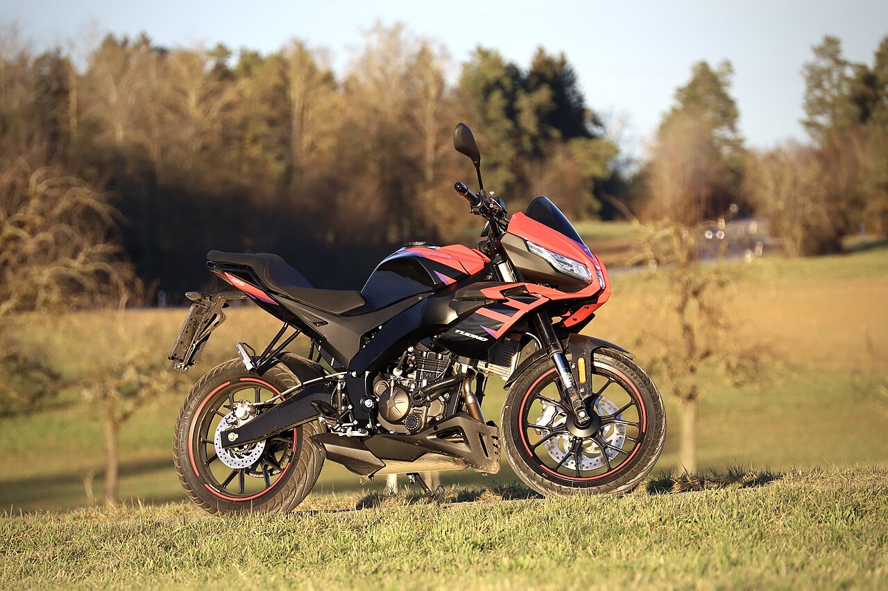 אופנוע Yamaha TMAX בצבע אדום-שחור, צילום צד מימין על דשא פתוח, רקע של שדה ועצים.