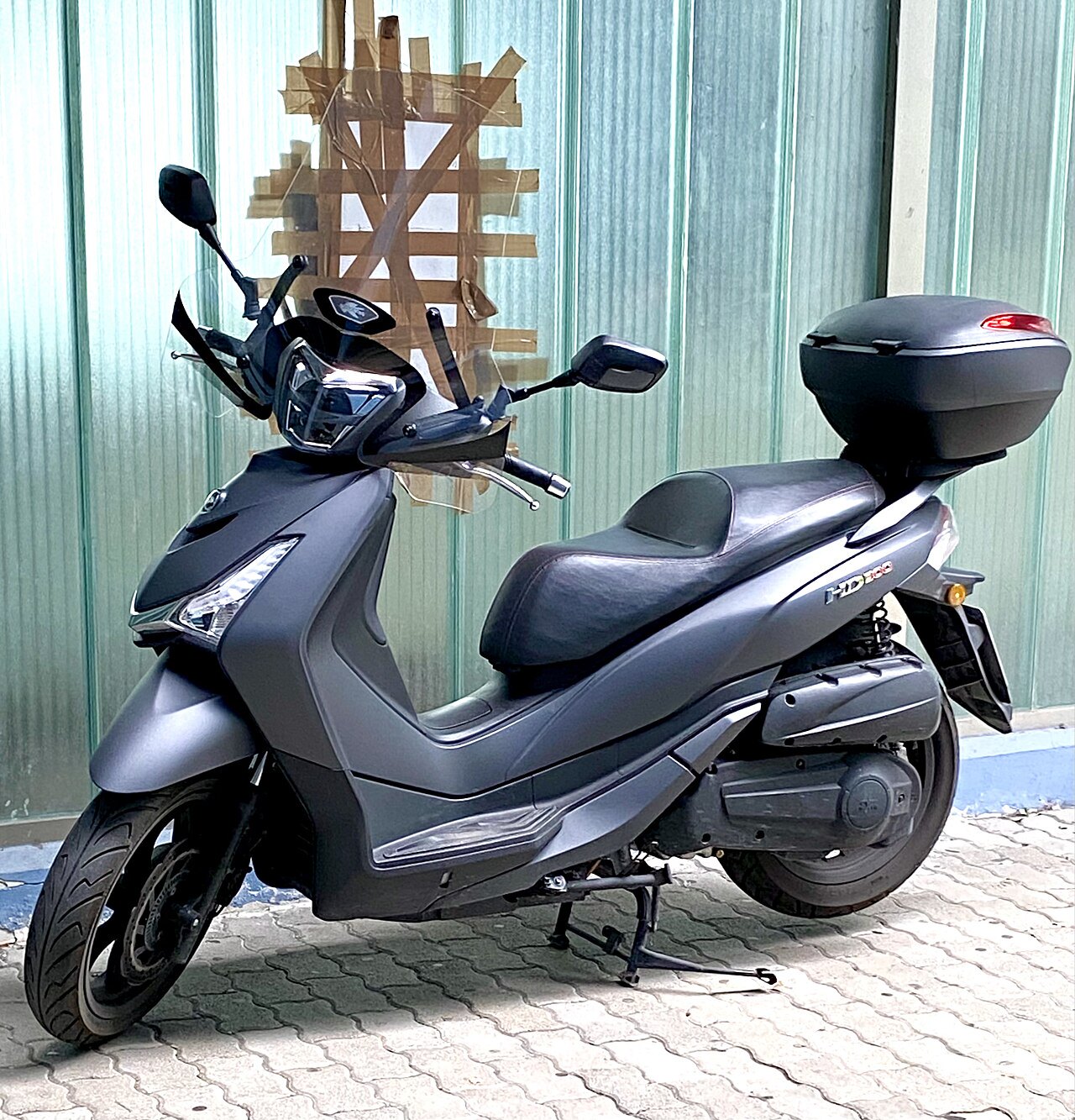 אופנוע חשמלי HD125 בצבע אפור מט, צילום צד, עם מארז אחורי, על רקע קיר מתכת ירוק-אפור