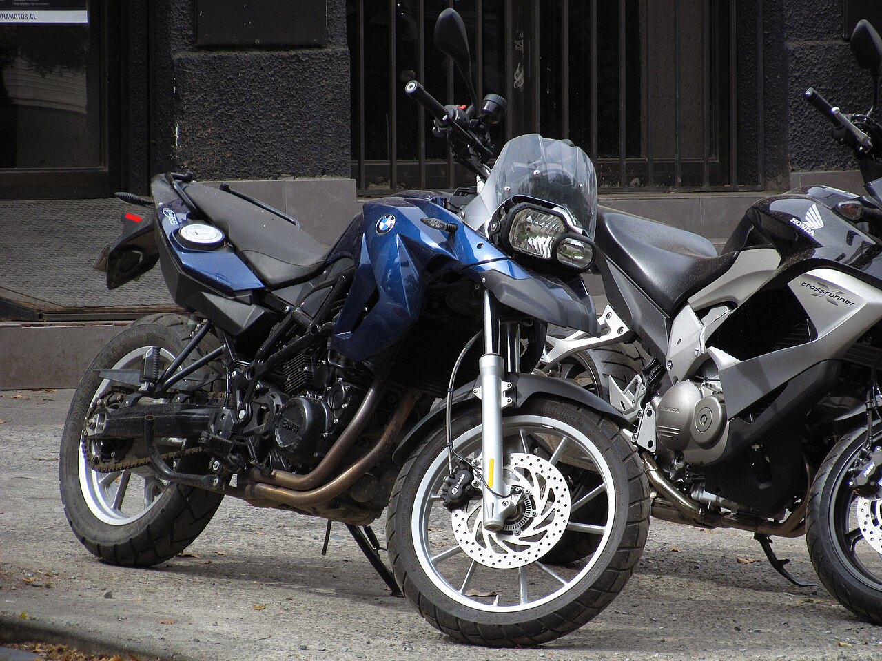 אופנוע BMW כחול עם מראה קדמי וגלגלים גדולים, עמיד על המדרכה לצד אופנוע Honda Crossrunner אפור, מול בניין עם חלונות מוגנים.