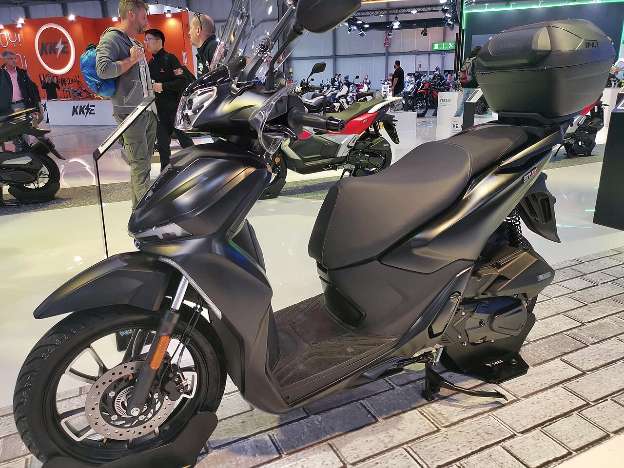 אופנוע חשמלי שחור מותג KEE, דגם GT, בתצוגה בתערוכה, זווית צדית עם מארז עליון, רקע של מבקרים וספקי אופנועים.