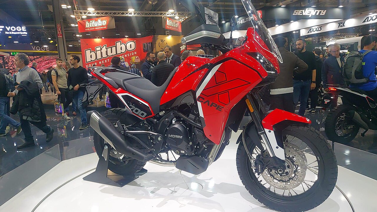 אופנוע מורייני Cape 700 אדום עם פלטפורמה לבנה בתערוכה, מבט צדדי, רקע של מבקרים וספקי ציוד