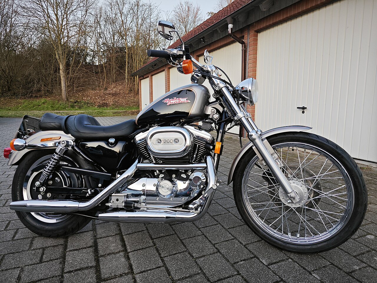 אופנוע Harley-Davidson 1200 בצבע שחור-כסף, צילום צד, על רצפת אבן מול גגון לבן, עצי אלון ברקע