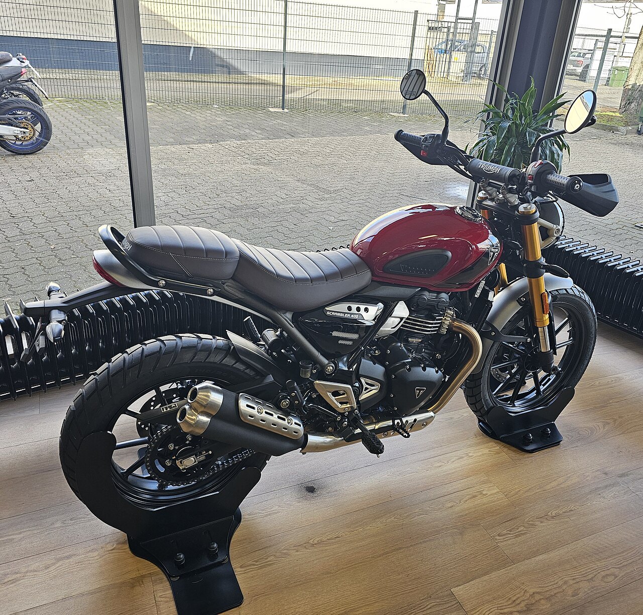 אופנוע Triumph Scrambler 400 אדום-שחור בתצוגה פנים, זווית צד, על רצפת עץ עם רקע חיצוני של גדר ופינת חנייה
