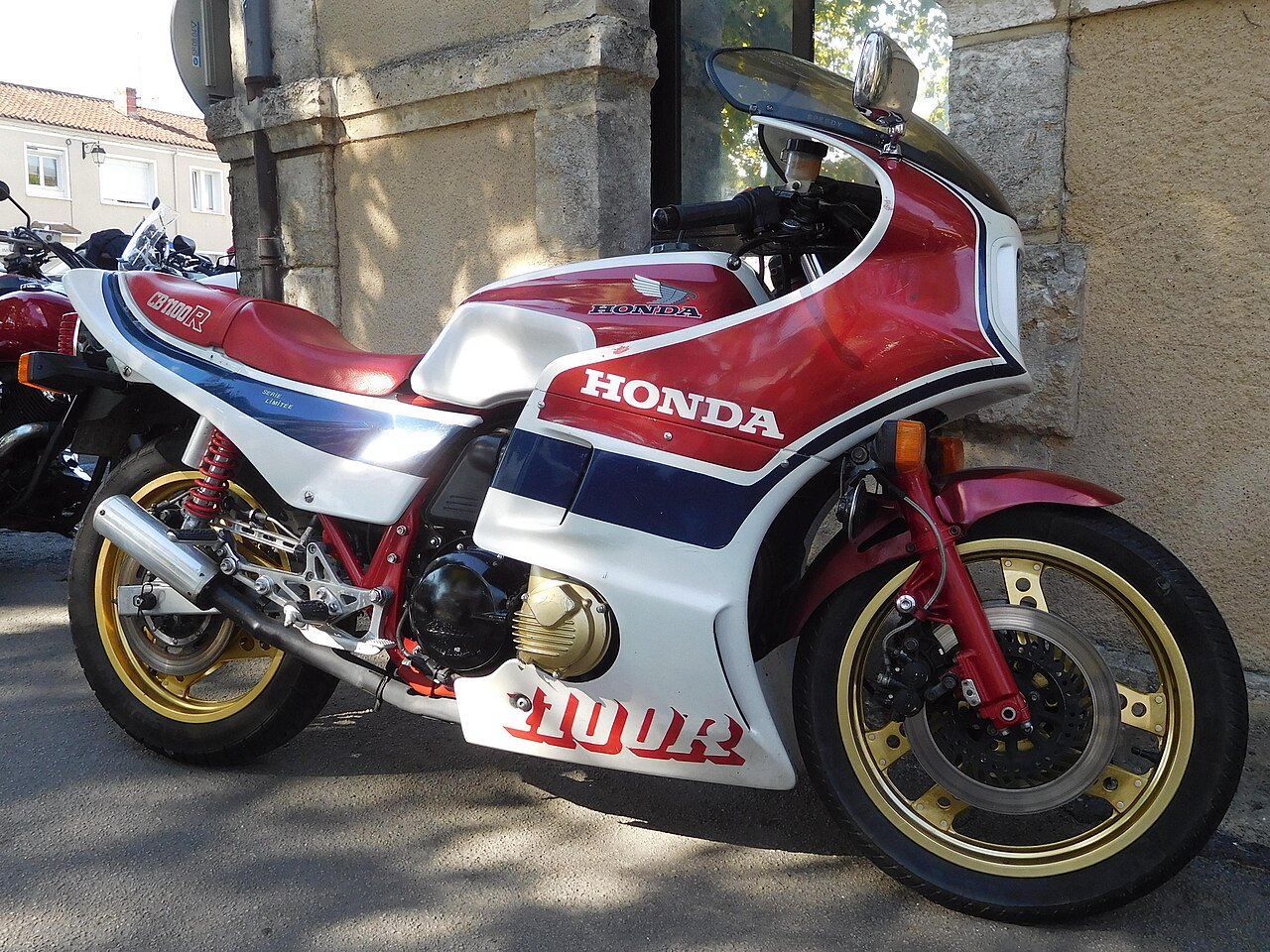 אופנוע הונדה CB1100R אדום-לבן-כחול, צילום צד, מונח מול קיר אבן, עם גלגלים זהובים וסמל HONDA בולט