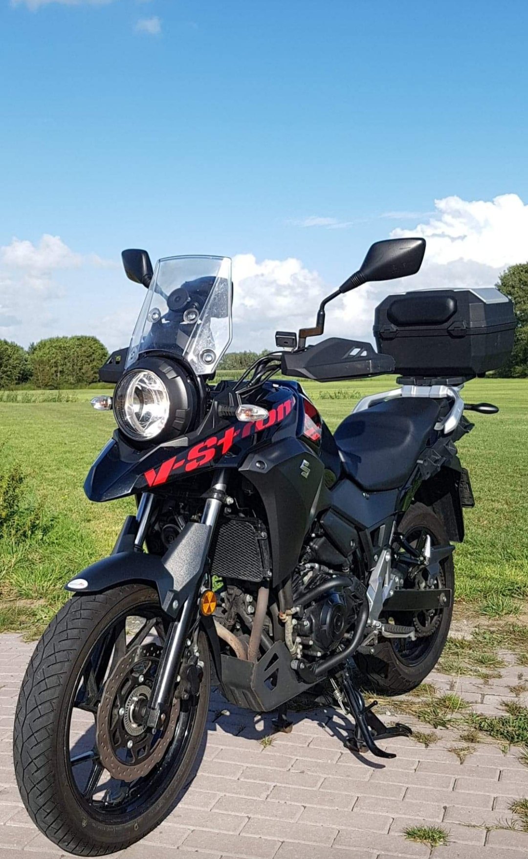 אופנוע Suzuki V-Strom שחור מוצב על אספלט בשדה ירוק, תחת שמיים כחולים עם עננים, זווית צילום קדמית-צדית.
