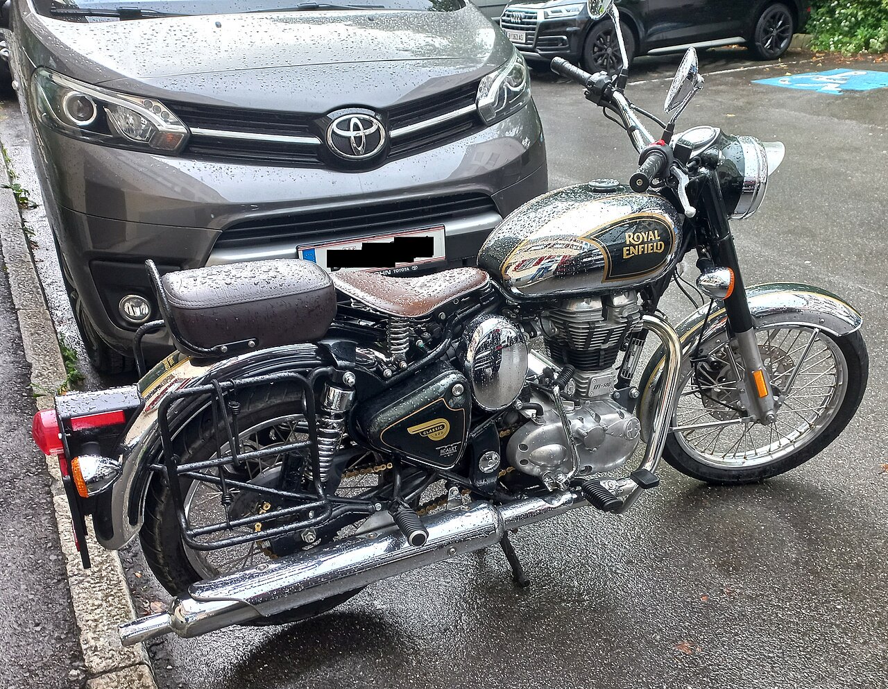 אופנוע ריאל אנפילד קלאסיק 350 שחור-כסוף עם מושב חום, מונח על חצובה רחבה, מים על הגוף, ברקע טויוטה אפור