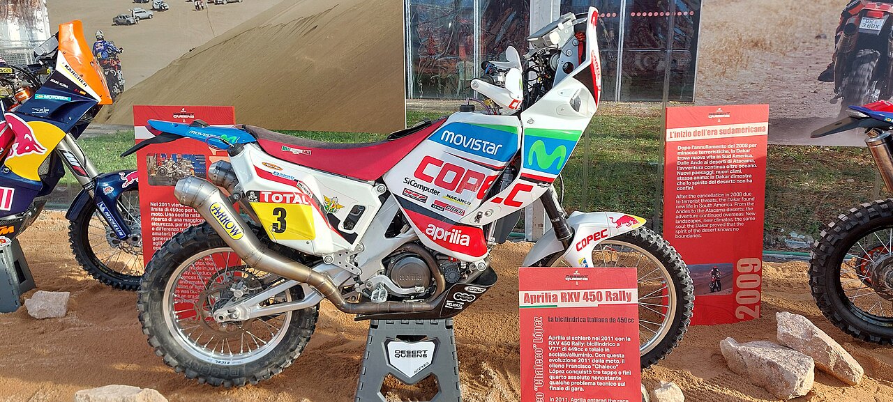 אופנוע Aprilia RSV 450 Rally לבן-אדום עם לוגו Movistar ו-COPEC, מוצג בתצוגה עם שלט 2009, רקע חול ורמלה.