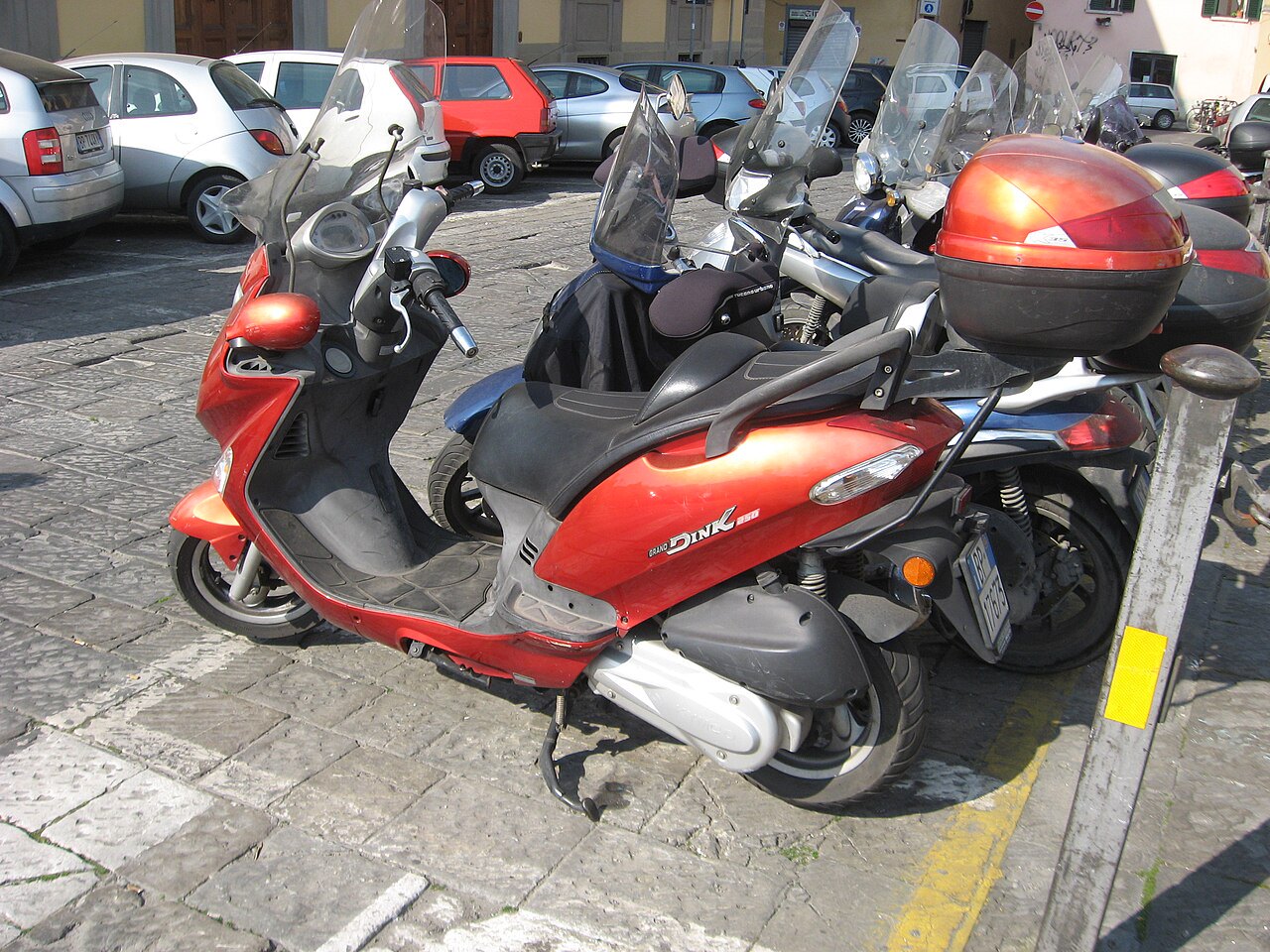 אופנוע סקוטר אדום דגם Grand Dink 250 חנייה על אספלט אבן, צד ימין, עם מנוע לבן ומסגרת שחורה, ברקע רכבים ורחוב עירוני.