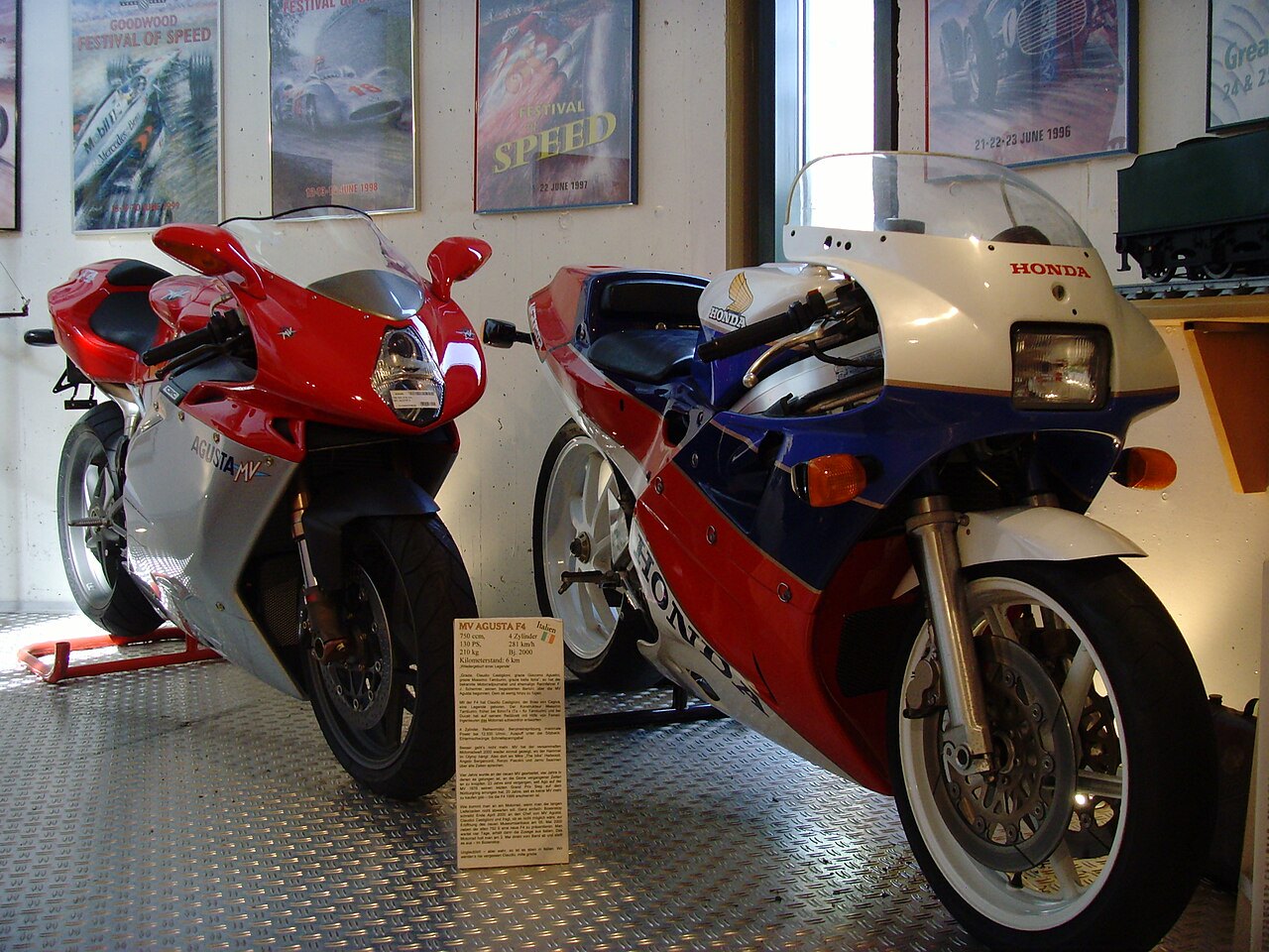 אופנועי ספורט MV Agusta אדום-כסוף ו-Honda לבן-כחול-אדום מוצגים בחלל מוזיאון, עם שלטים של פסטיבל מהירות ברקע