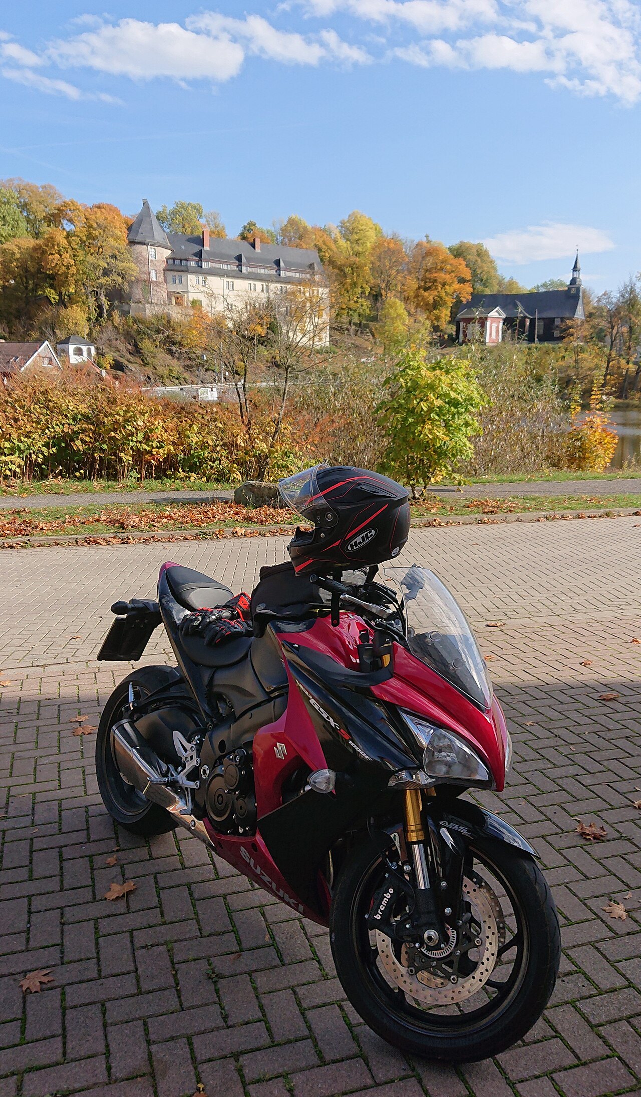 אופנוע סוזוקי GSX-S1000 אדום-שחור עמיד על שטח אבן, עם קסדה HJC על הידית, ברקע חורש אוטמף ומבנה עתיק על תל