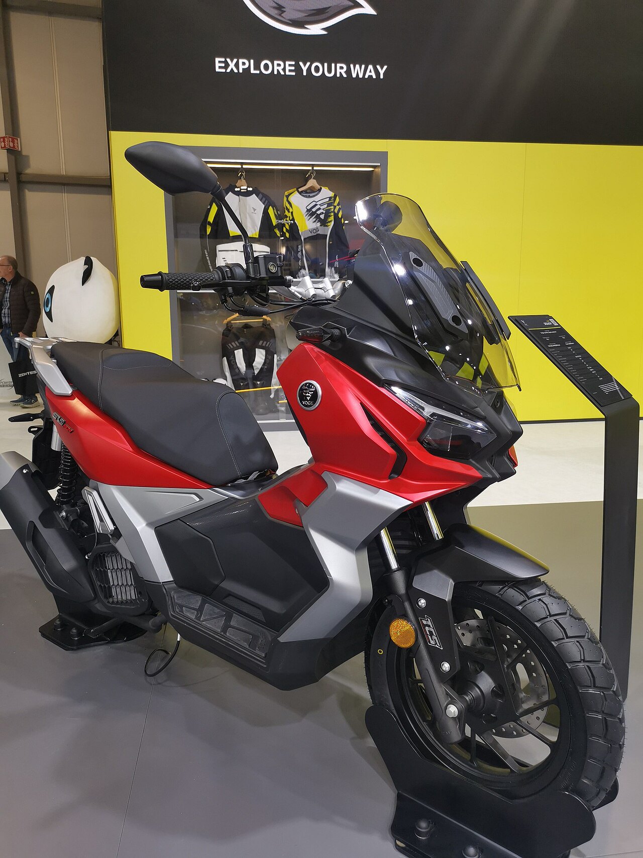 אופנוע Voge S160T אדום-שחור עם מראה ענק, צילום זווית צד-קדימה בפנימיות תערוכה עם רקע צהוב וטקסט 'EXPLORE YOUR WAY'