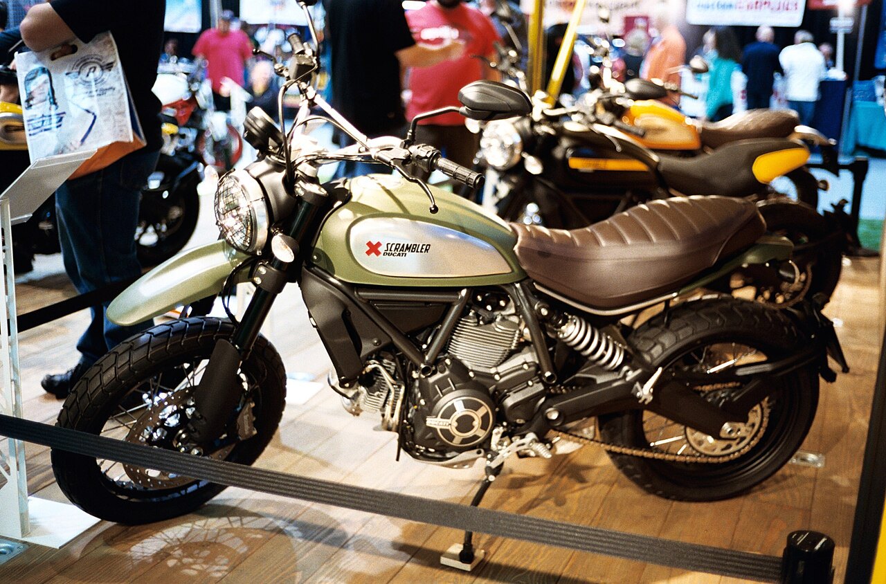 אופנוע Ducati Scrambler בצבע ירוק-זהב, צילום צד, בתצוגה עם אנשים ברקע