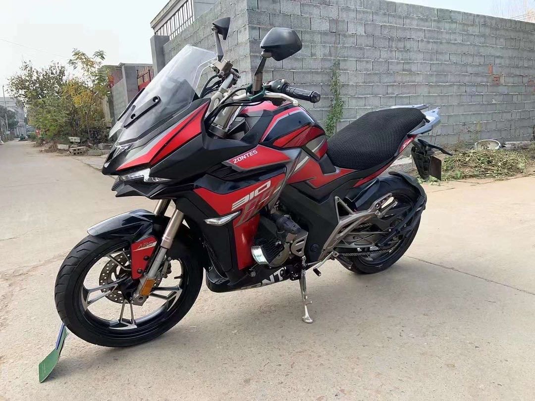 אופנוע Zontes 310 אדום-שחור, צילום צד, עמיד על רצפת בטון מול חומה אבן, עם מראה ומסך עליון.