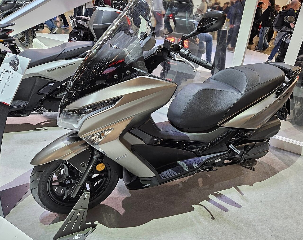 אופנוע קיימקו X-TOWN 300 בצבע חום-כסוף, צילום צד קרוב בזווית נמוכה, בתצוגה פנימית עם אנשים ברקע.