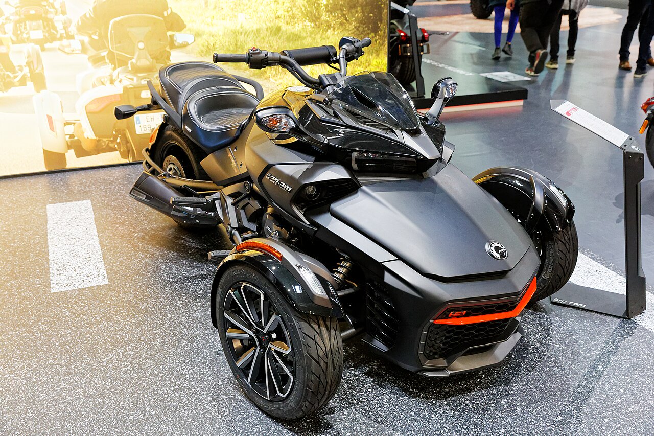 אופנוע תלת-אופן Can-Am סלבריטי אפור עם פס אדום בקדמת, צילום זווית גבוהה על שטח תצוגה, רקע עם אנשים ומודלים נוספים