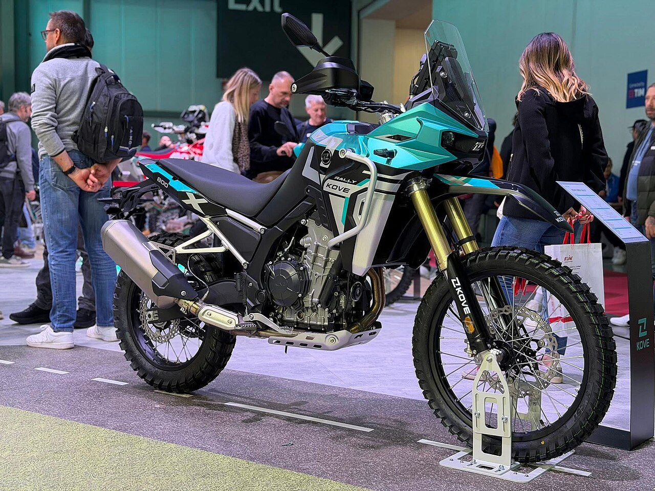 אופנוע אדベンчер ZKOVE Rally 600 בצבע טורקיז וסגול, צולם מזווית צדית בפנימיות תערוכה עם מבקרים ברקע.