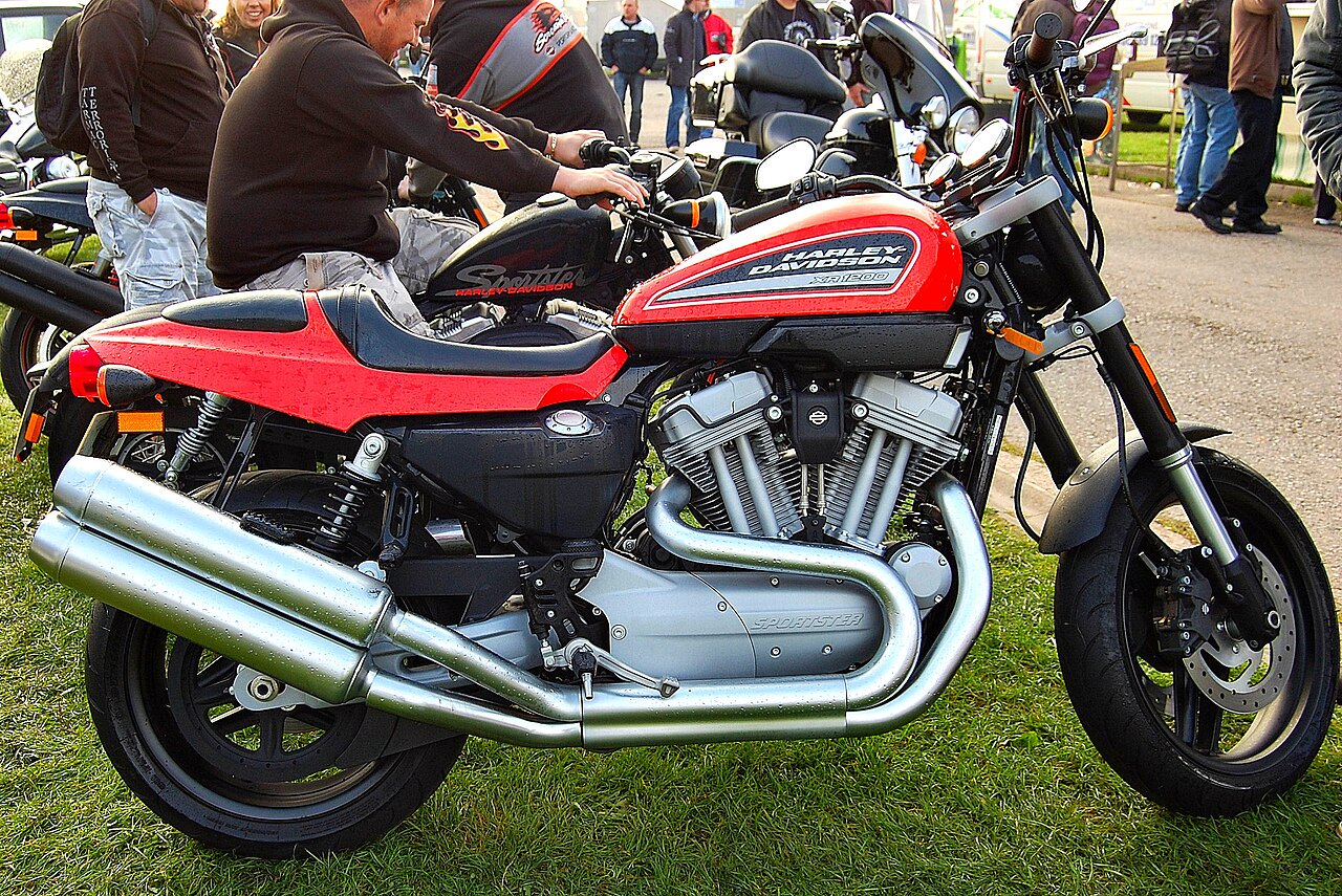 אופנוע Harley-Davidson XR1200 אדום-שחור, צילום צד, על דשא, עם מנוע V-Twin ומערכת עזרות מטאליות, בסביבה של מפגש אופנוענים.