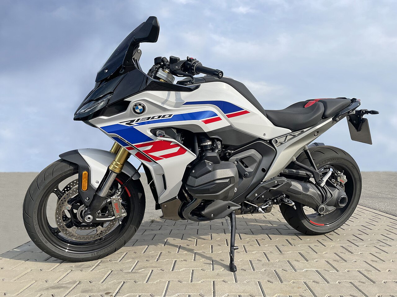 אופנוע BMW R1200R לבן עם פסים כחולים ואדומים, צילום צד, על מדרכה בזווית נמוכה, רקע שמיים כחולים עם עננים