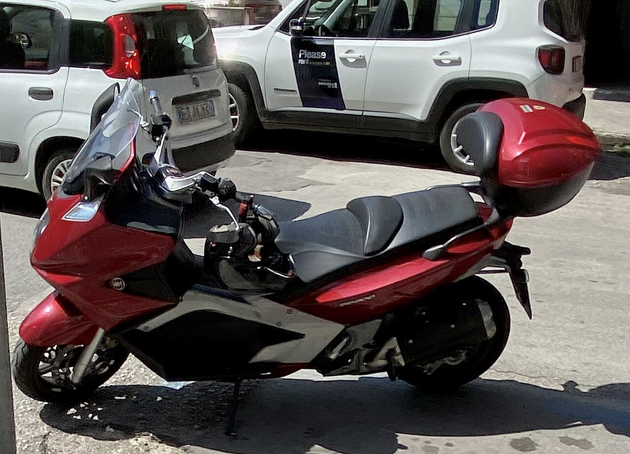 אופנוע אדום וכסוף מותג Honda, דגם Forza, מונח על רצפת בטון עם תיק אחורית אדום, ברקע רכבים לבנים.