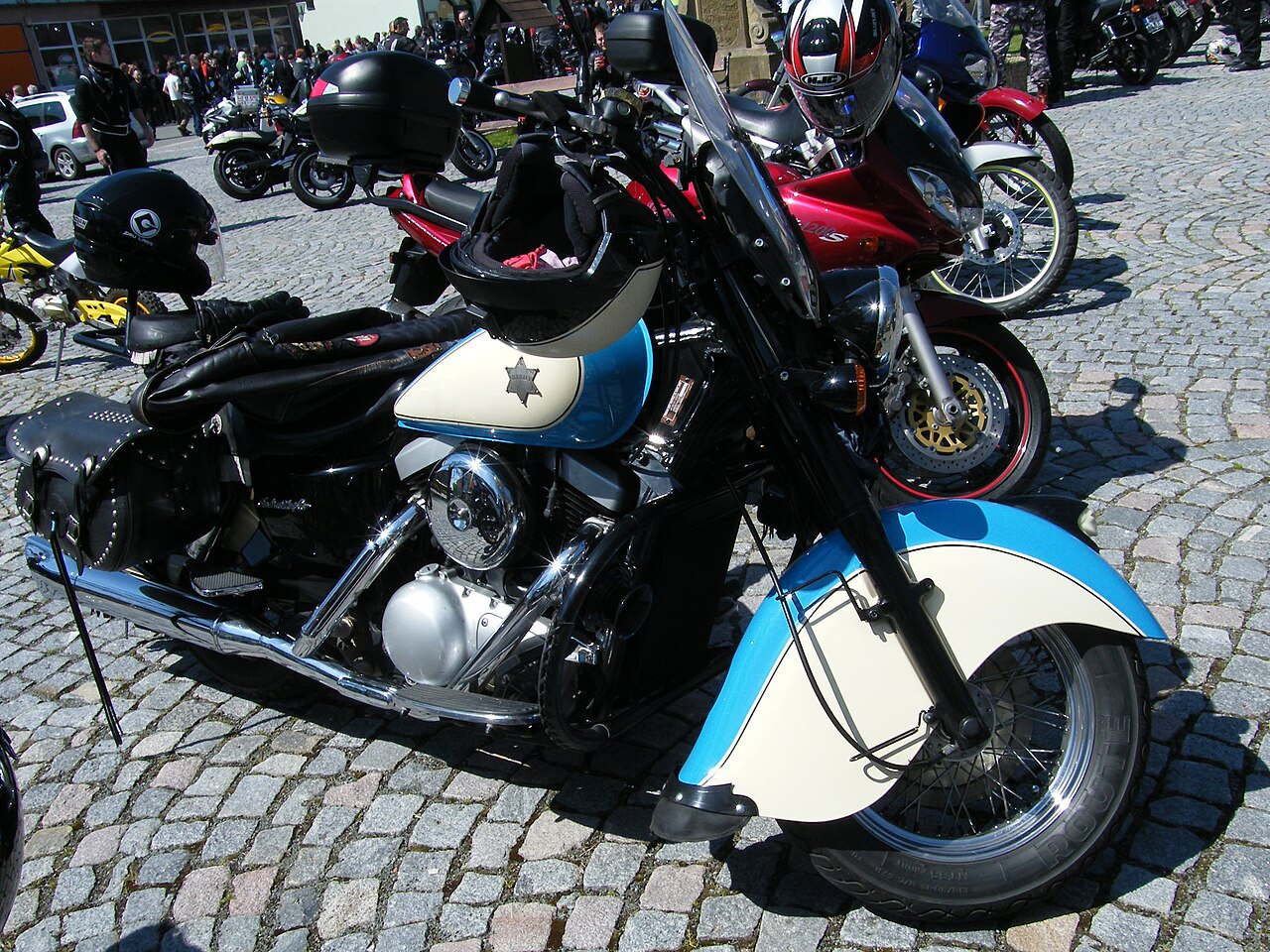 אופנוע Harley-Davidson קלאסי בצבע כחול-לבן עם מושב שחור וסנדליים, צולם מזווית קדמית על רצפת אבן, עם אופנועים נוספים ברקע.