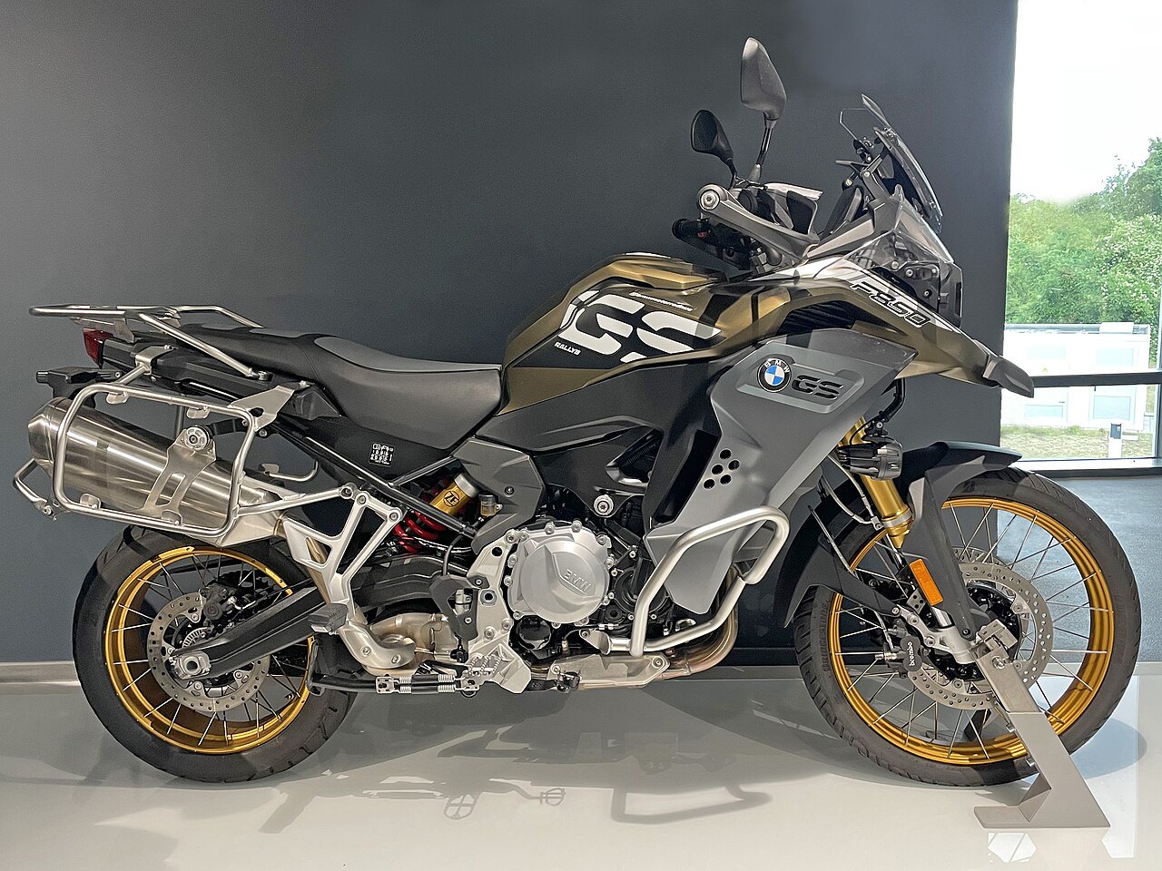 אופנוע BMW R 1300 GS בצבע אפור-זהב, צילום צד, מוצג בחלל מודרני עם חלון ברקע, כולל מוגן מנוע ותאורה LED