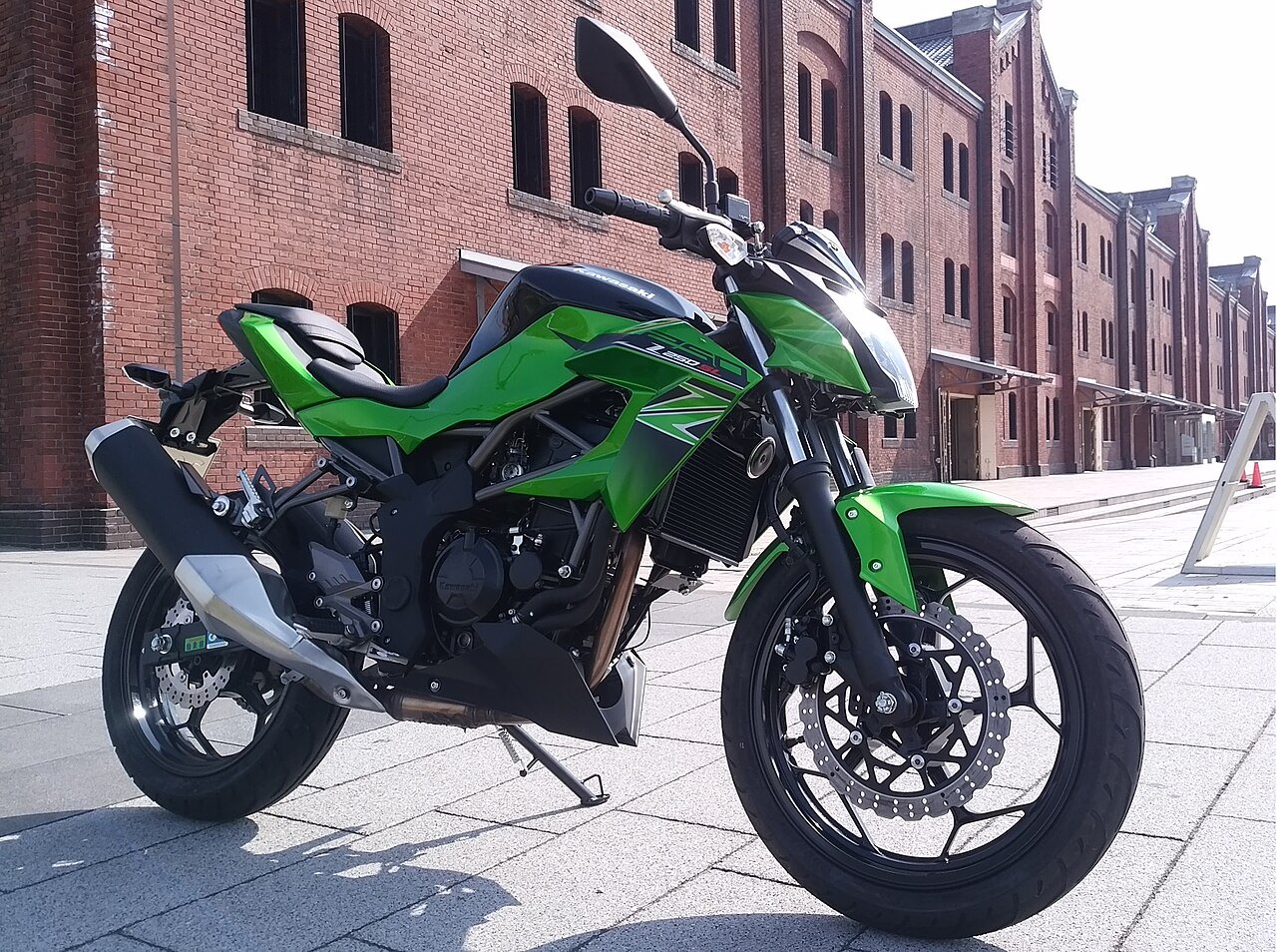 אופנוע Kawasaki Z650 בצבע ירוק בהיר, צילום צד מקרוב על רקע בניין אדום, מוצג על מדרכה בזווית שמש.
