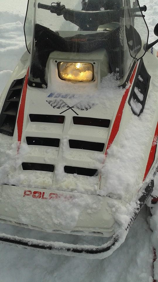אופנוע שלג לבן עם פסים אדומים, מותג POLAR, צילום קרוב מלפנים, מואר, מוסתר חלקית בשלג, על רקע שלג.