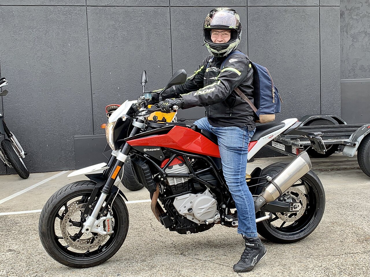 אופנוע Husqvarna אדום-שחור עם רוכב בלבוש אבטחה, צילום מזווית צדית מול קיר אפור, מותג ורוכב בלבוש מקצועי